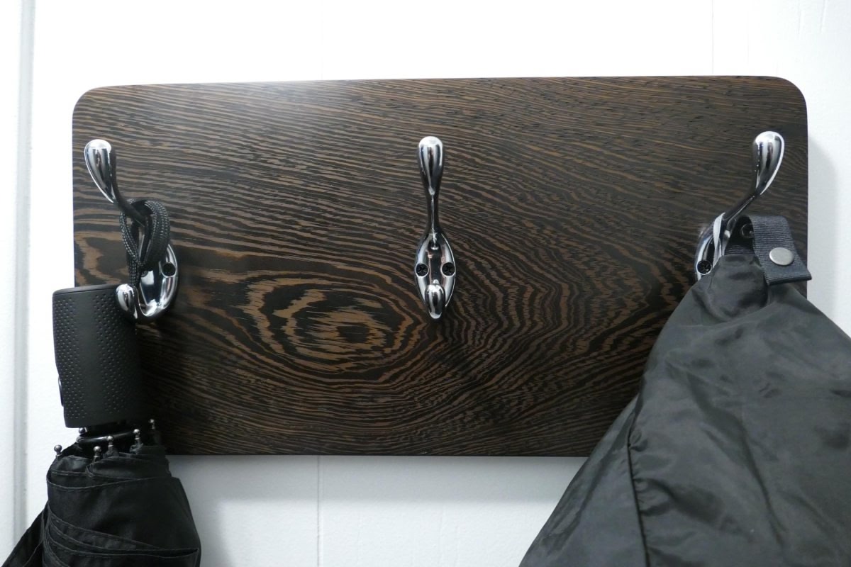 A black and tan wenge hardwood coat rack with 3 chrome coat hooks hanging on a white wall