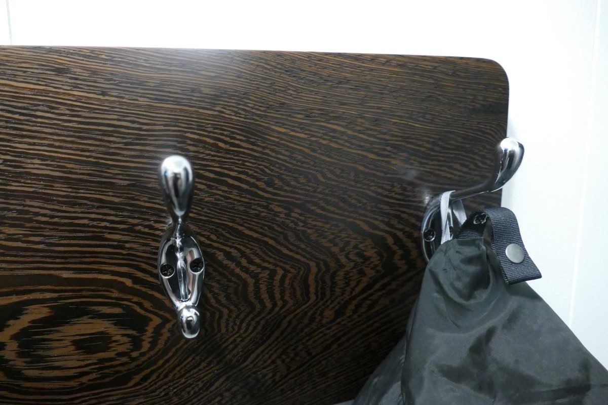A black and tan wenge hardwood coat rack with 3 chrome coat hooks hanging on a white wall
