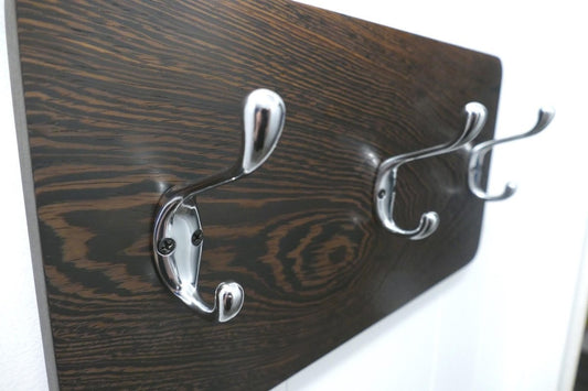 A black and tan wenge hardwood coat rack with 3 chrome coat hooks hanging on a white wall