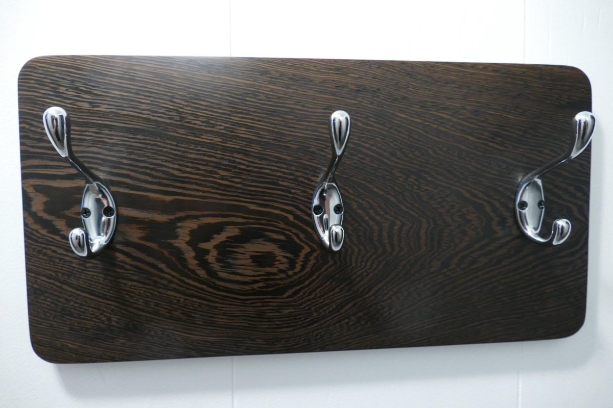A black and tan wenge hardwood coat rack with 3 chrome coat hooks hanging on a white wall