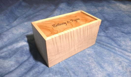 A soft curly maple wood box with an engraved curly cherry sliding lid and a curly walnut bottom sitting on a blue background
