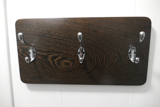 A black and tan wenge hardwood coat rack with 3 chrome coat hooks hanging on a white wall