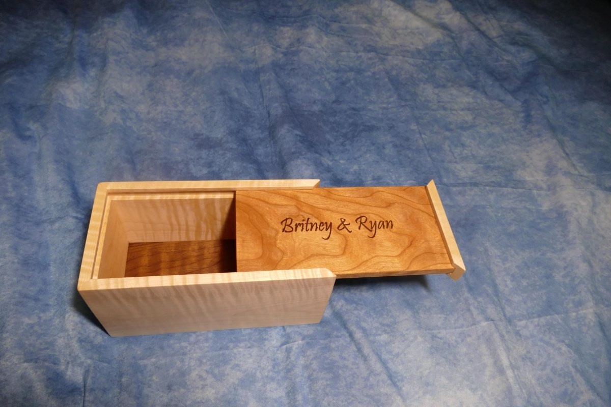 A soft curly maple wood box with an engraved curly cherry sliding lid and a curly walnut bottom sitting on a blue background
