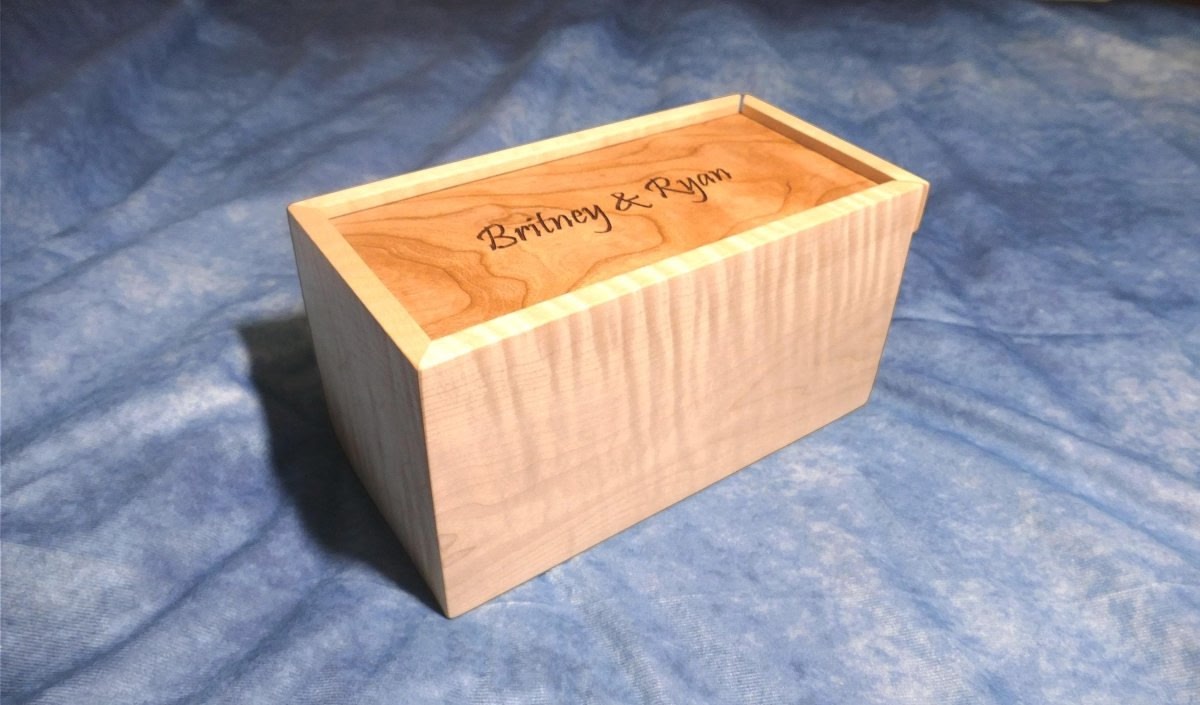 A soft curly maple wood box with an engraved curly cherry sliding lid and a curly walnut bottom sitting on a blue background